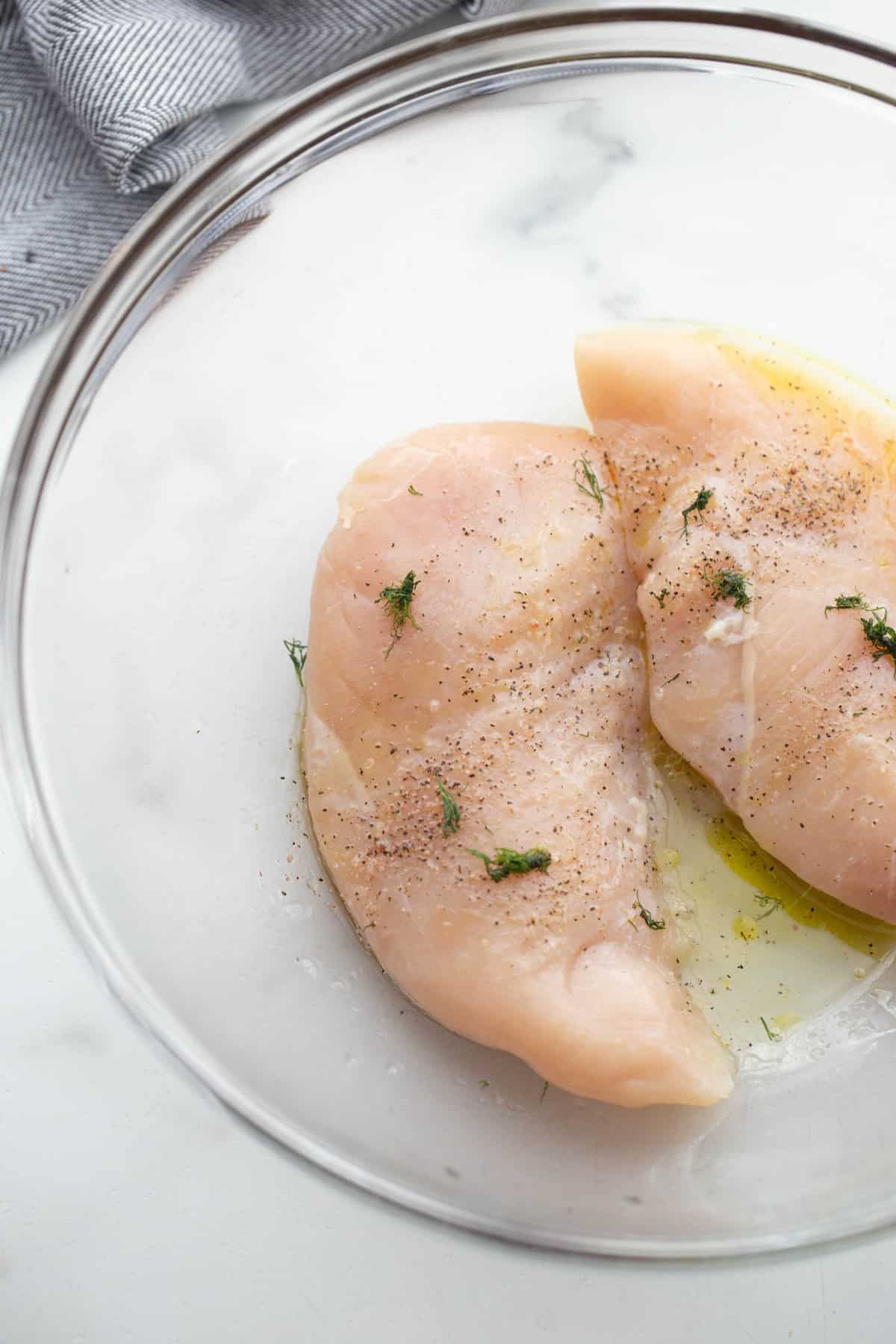 Raw chicken breast in a bowl with lemon, olive oil, dill, sat, and pepper.