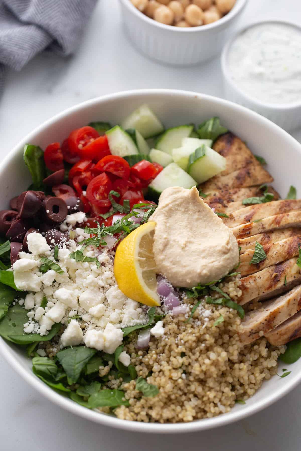 Medium white bowl filled with chicken, quinoa, feta, veggies, green, hummus and garnished with a lemon.