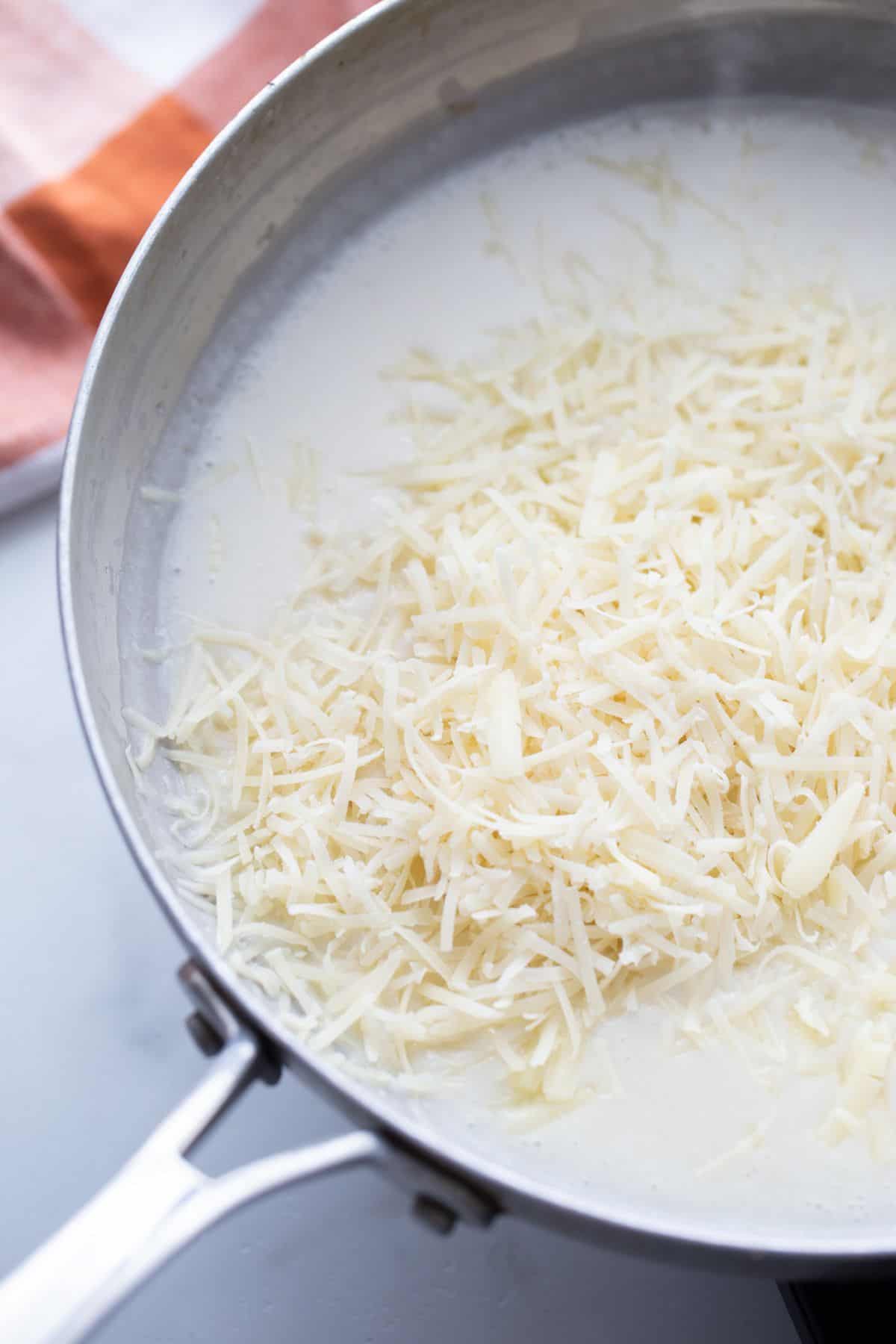 Shredded cheese being melted in a pan of healthy alfredo sauce.