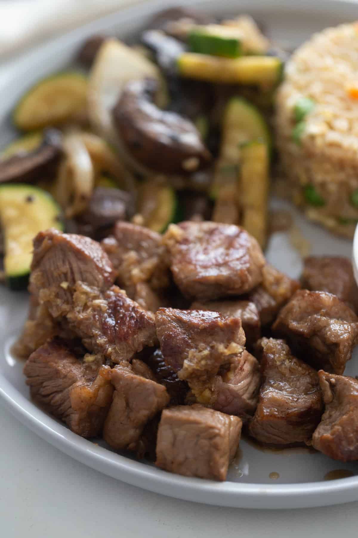 Cubed steak drizzled with ginger sauce on a plate; sauteed veggies and rice are on the same plate in the background, out of focus