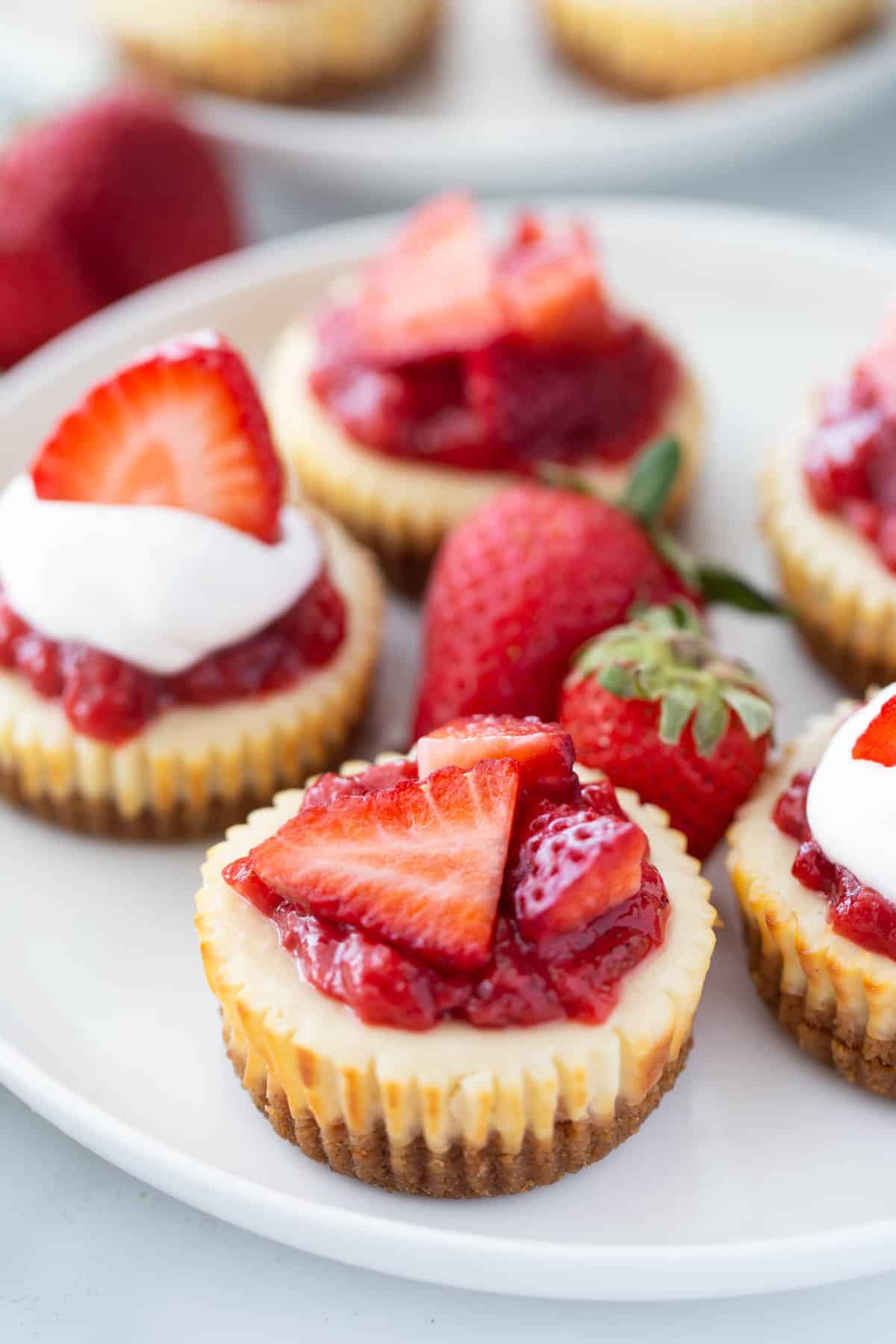 Close up image of Healthy Mini Cheesecakes with strawberry topping on a white plate. The ones in front are in focus, and the ones at the top of the image are out of focus.