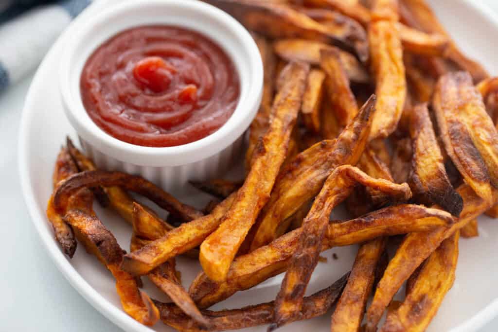 Plate of crispy, golden brown air fryer sweet potato fries next to a ramekin of ketchup.