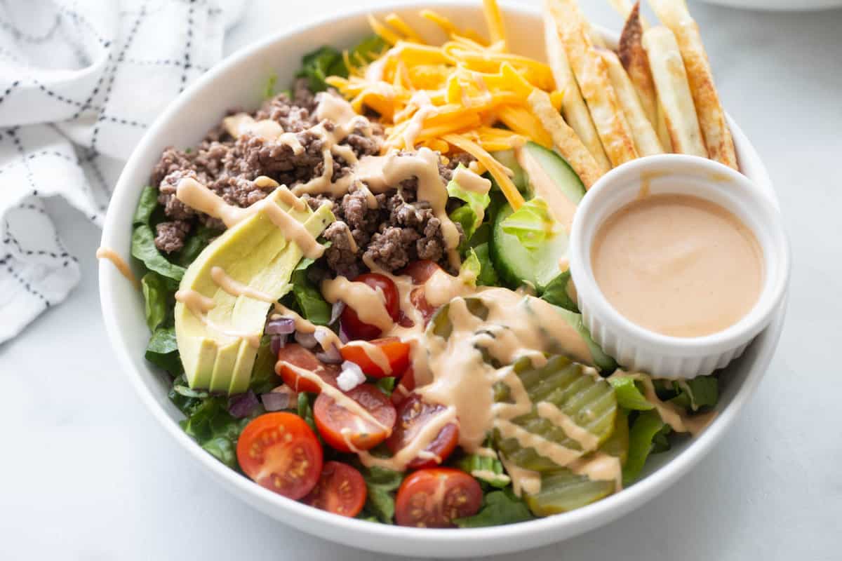 Burger bowl with ground beef, tomatoes, pickles, lettuce, sliced avocado, fries, and a side of special sauce.