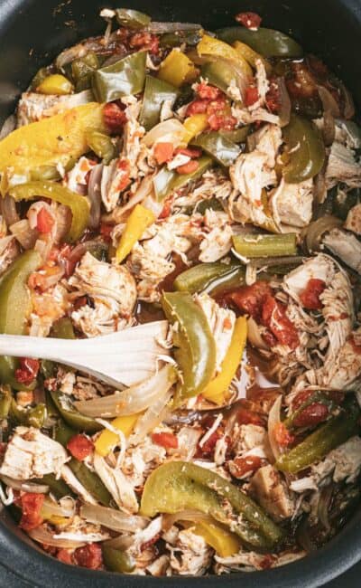 An oval shaped black slow cooker with shredded chicken and fajita veggies with a wooden spoon placed inside.