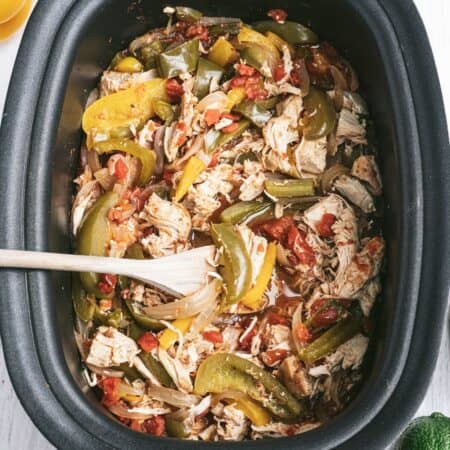 An oval shaped black slow cooker with shredded chicken and fajita veggies with a wooden spoon placed inside.