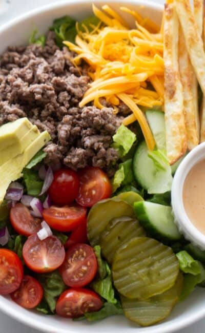 Burger bowl with ground beef, tomatoes, pickles, lettuce, sliced avocado, fries, and a side of special sauce.