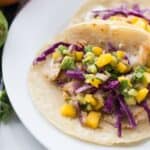 Two Fish Tacos with Mango Salsa and red cabbage on corn tortillas on a white plate.