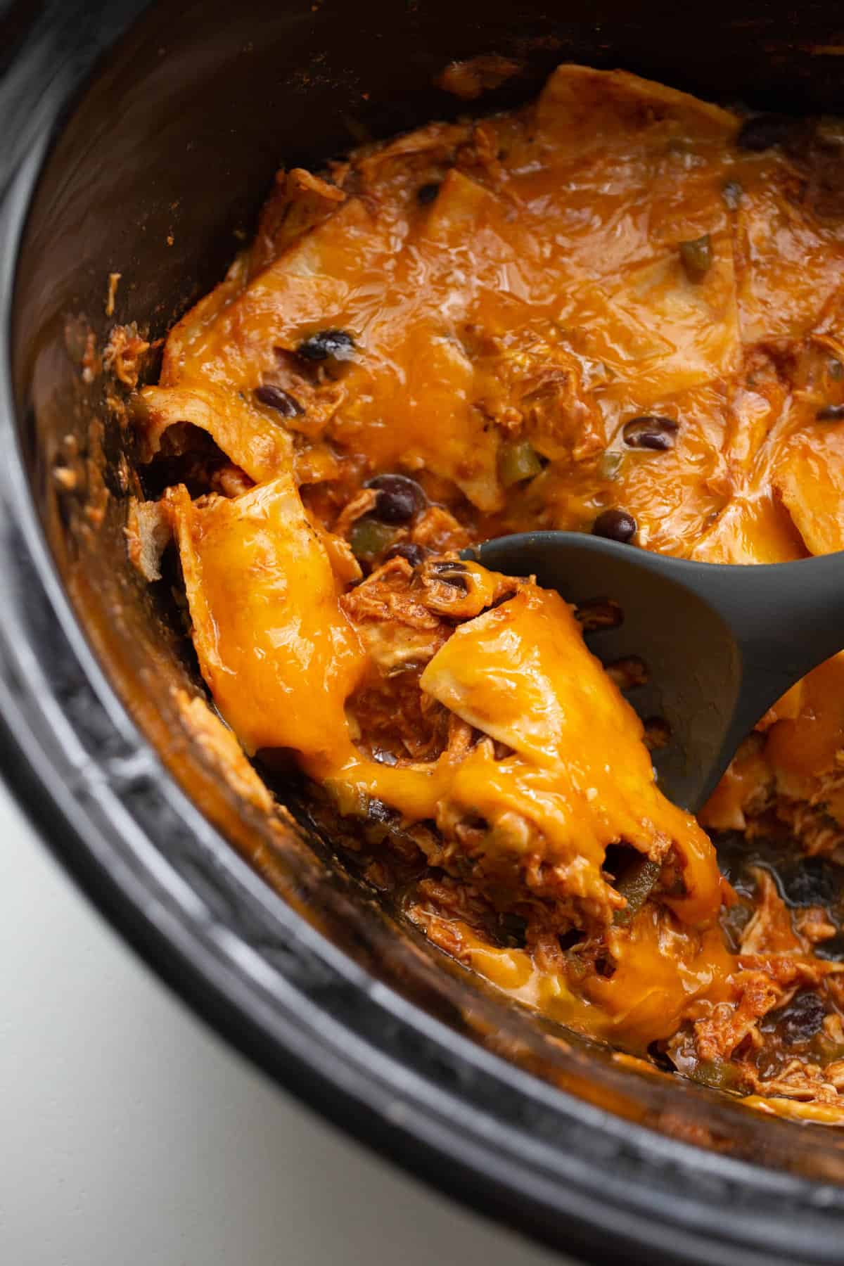Chicken enchilada casserole in a slow cooker with a black spoon scooping out a portion of cheesy goodness.