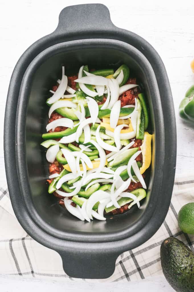 An oval shaped black slow cooker with raw chicken and fajita veggies.