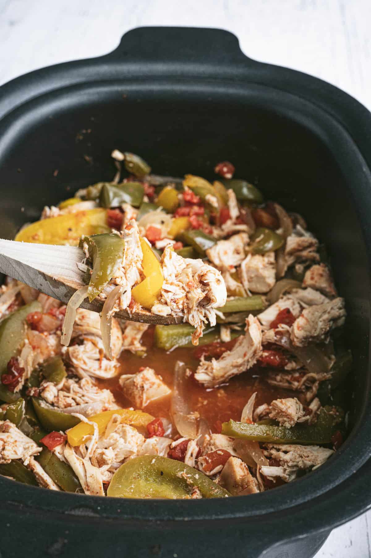 An oval shaped black slow cooker with shredded chicken and fajita veggies with a wooden spoon scooping up a portion.