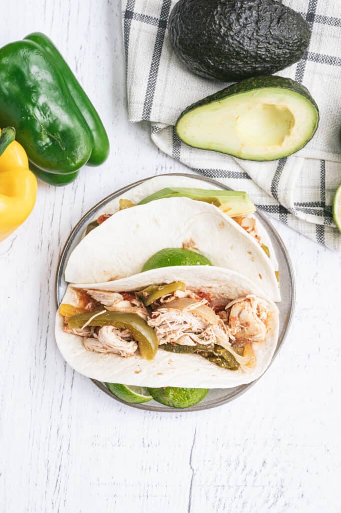 Chicken fajitas inside white corn tortillas on a white wood background with bell peppers and avocados sitting nearby.