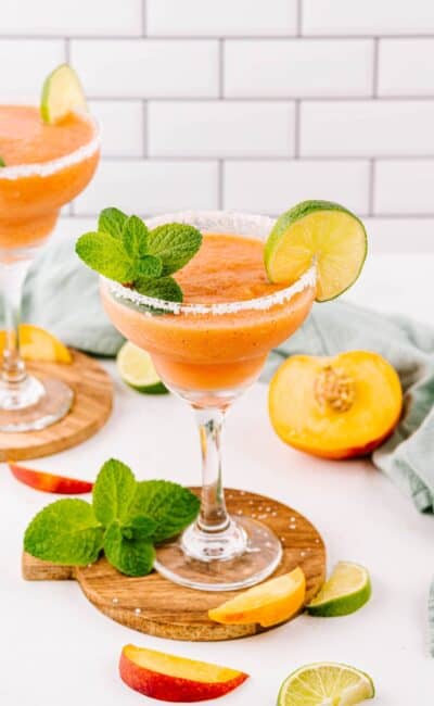 Salt rimmed frozen peach margarita cocktails on a white table with limes and peach slices styled around the drinks.