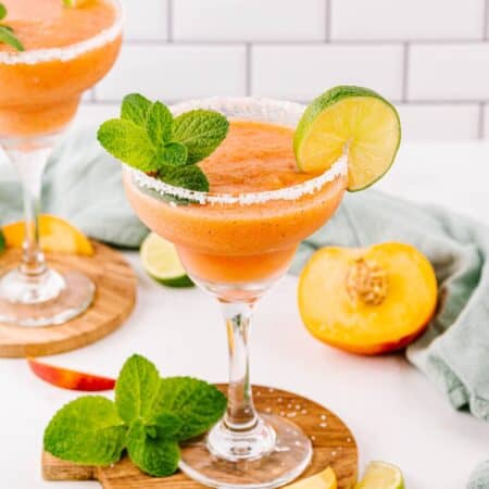 Salt rimmed frozen peach margarita cocktails on a white table with limes and peach slices styled around the drinks.