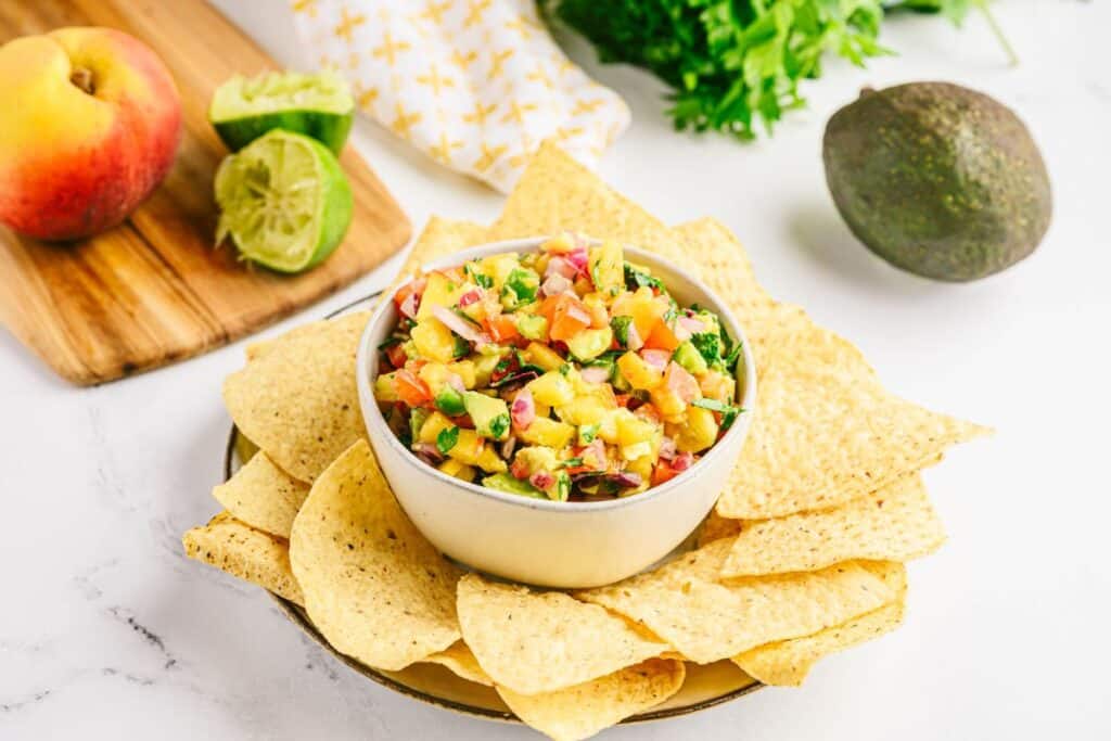 Homemade fresh peach salsa in a small bowl surrounded by tortilla chips, an avocado, lime, and a peach.