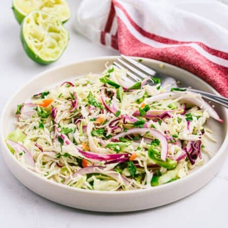 A white dish of cilantro lime coleslaw plated with a fork and a red striped fabric napkin nearby.