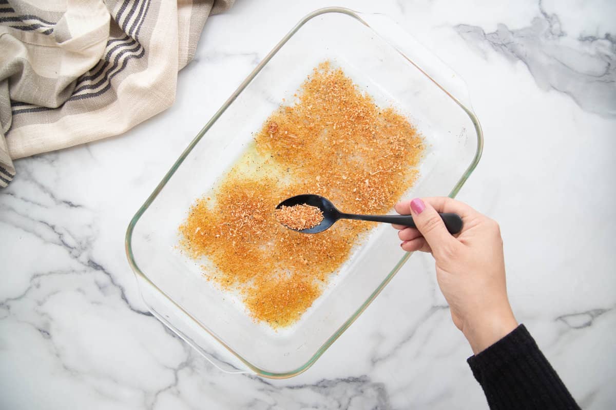 Sprinkling parmesan cheese and spices over olive oil in a glass baking dish.