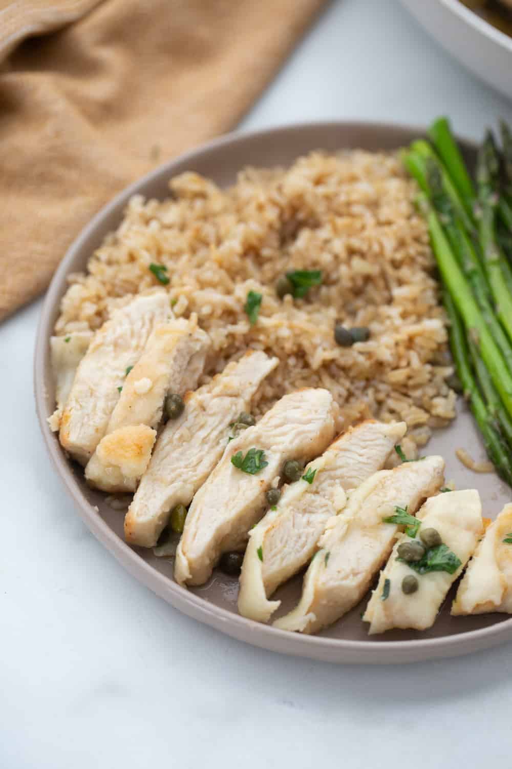 Chicken Scallopini served with roasted asparagus and brown rice shown sliced on a neutral colored rimmed plate.