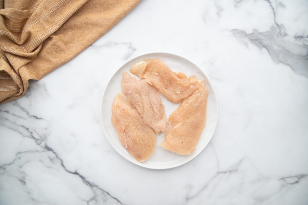 Thinly sliced lightly seasoned chicken breasts on a white plate on a white marble countertop.