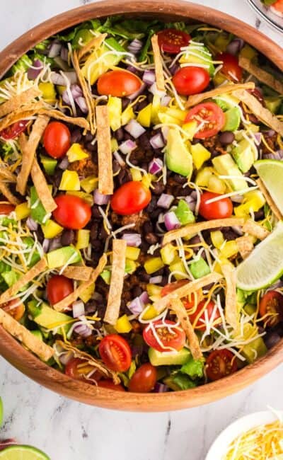 Healthy Tacos Salads in a wooden bowl topped with tortilla strips, cheese, avocado, and fresh lime.