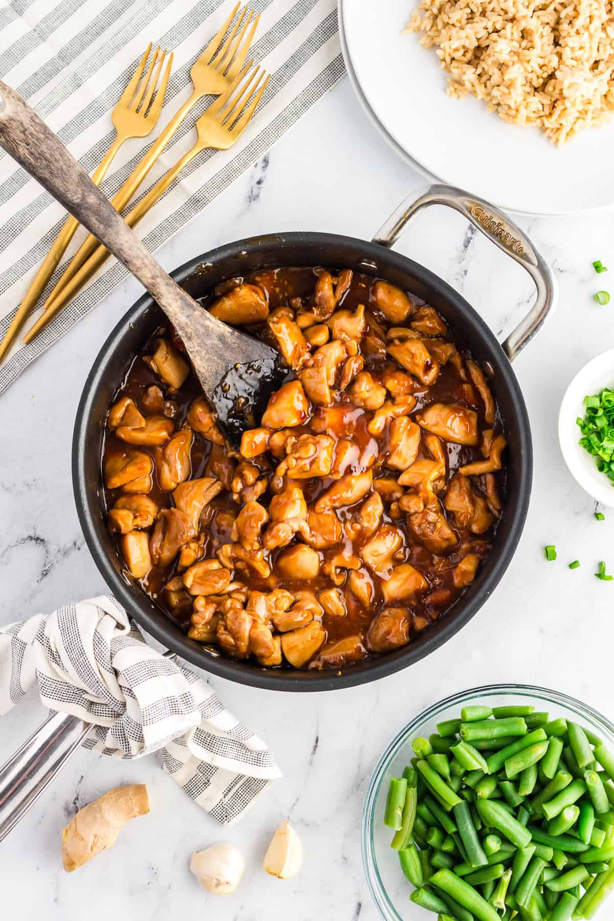 A skillet of homemade bourbon chicken with a brown rice, green beans, and a small bowl of green onions sitting to the side.