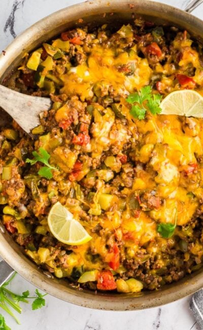 Overhead view of a colorful taco zucchini skillet in a pan, topped with melted cheese and sliced avocado, with lime wedges.