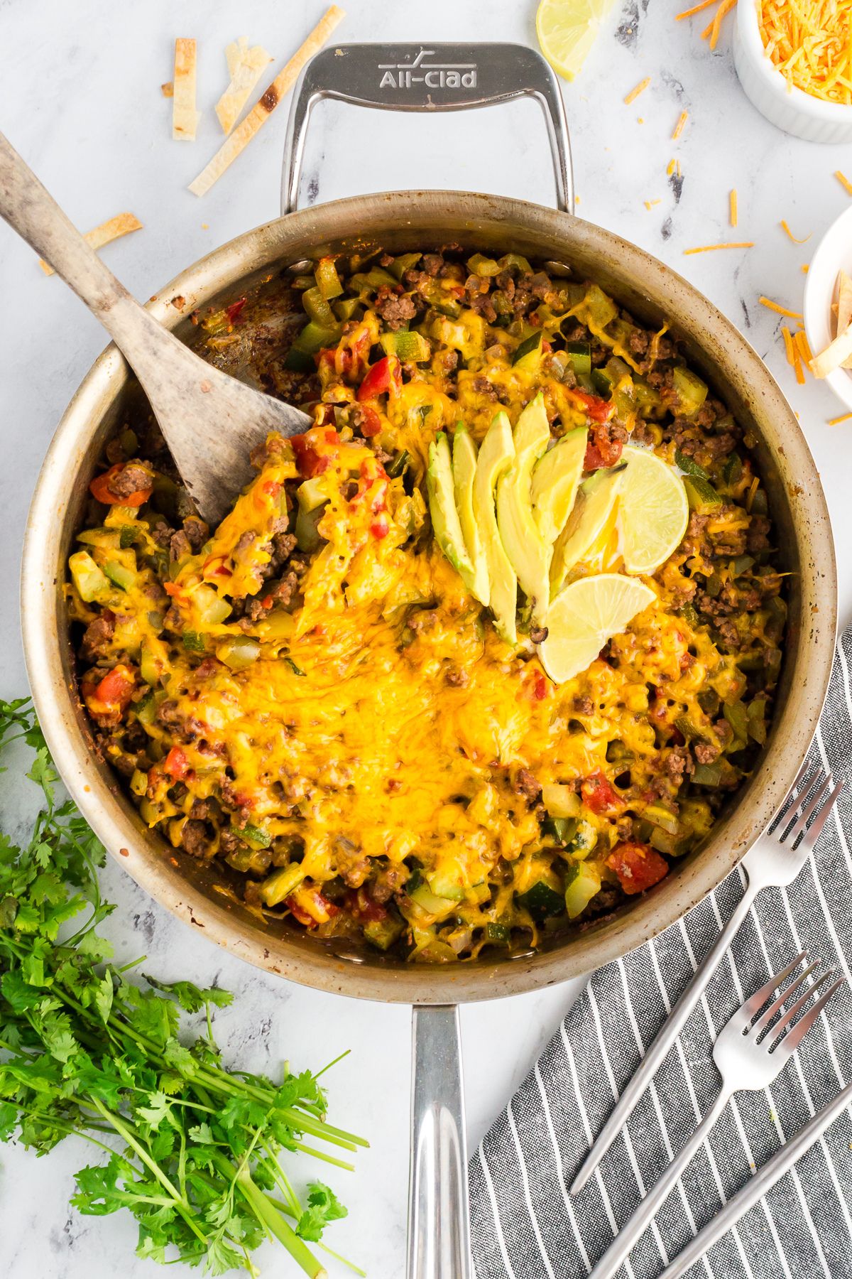 Overhead view of a colorful taco zucchini skillet in a pan, topped with melted cheese and sliced avocado, with lime wedges.