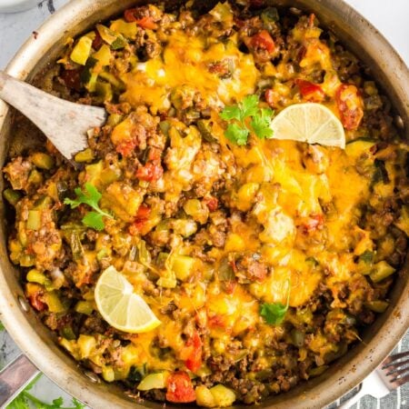 Overhead view of a colorful taco zucchini skillet in a pan, topped with melted cheese and sliced avocado, with lime wedges.