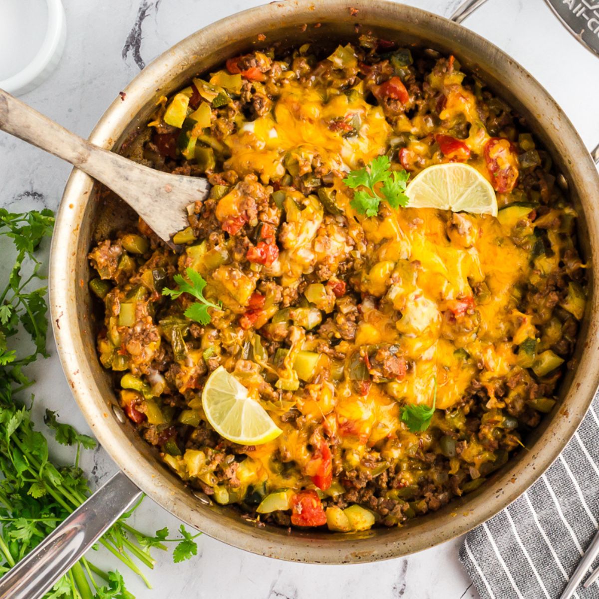 Overhead view of a colorful taco zucchini skillet in a pan, topped with melted cheese and sliced avocado, with lime wedges.