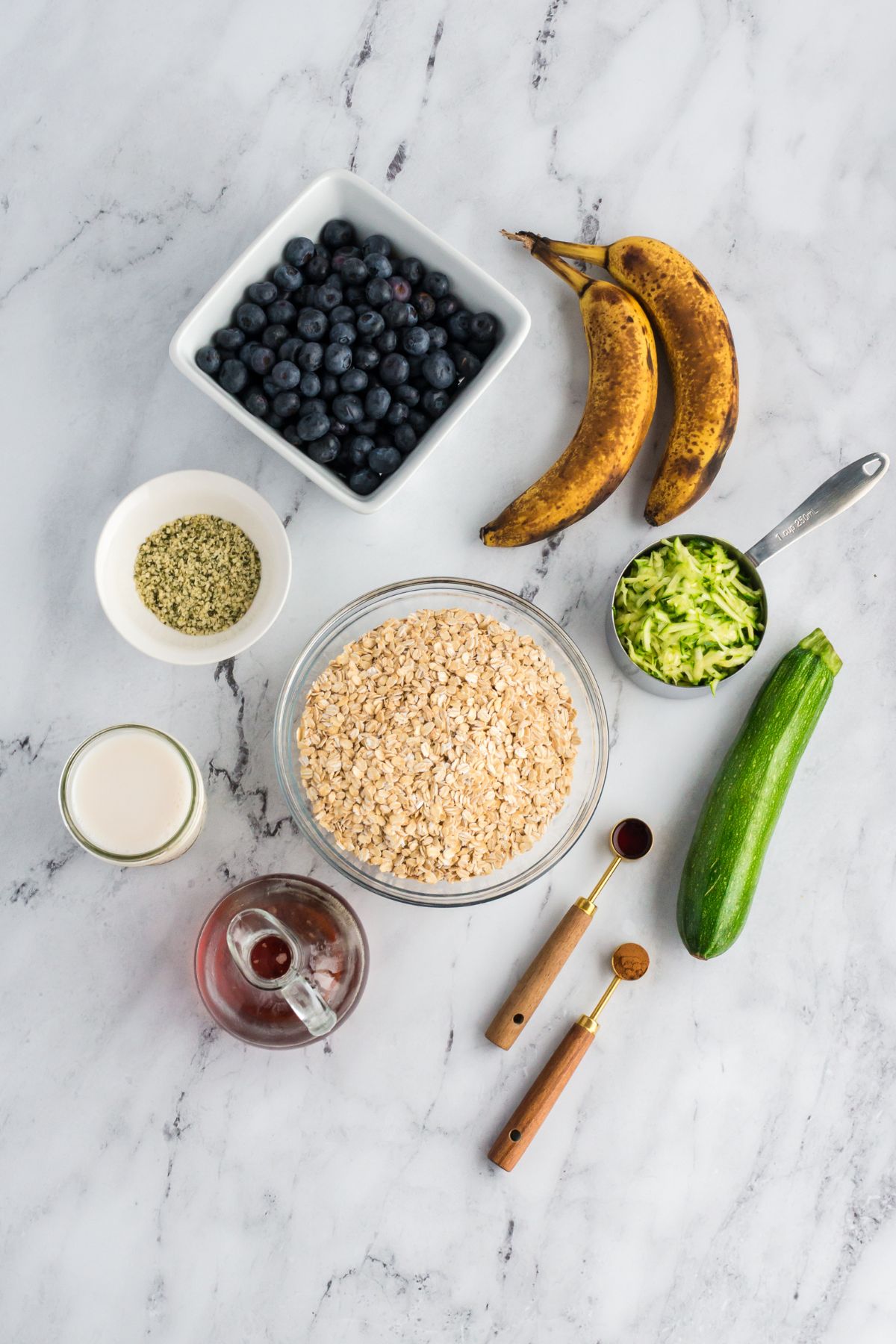 Ingredients for zucchini blueberry baked oatmeal including oats, blueberries, bananas, zucchini, milk, and spices arranged on a marble surface.