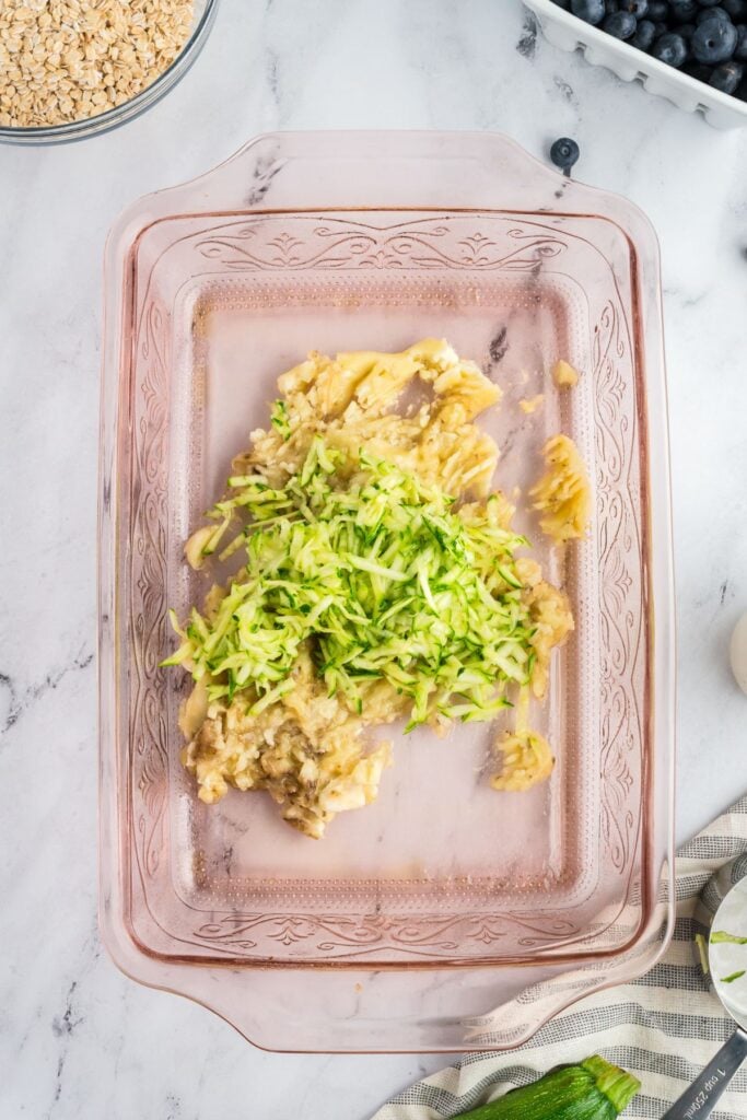 A glass baking dish with mashed bananas and shredded zucchini, ready to be mixed with other ingredients.