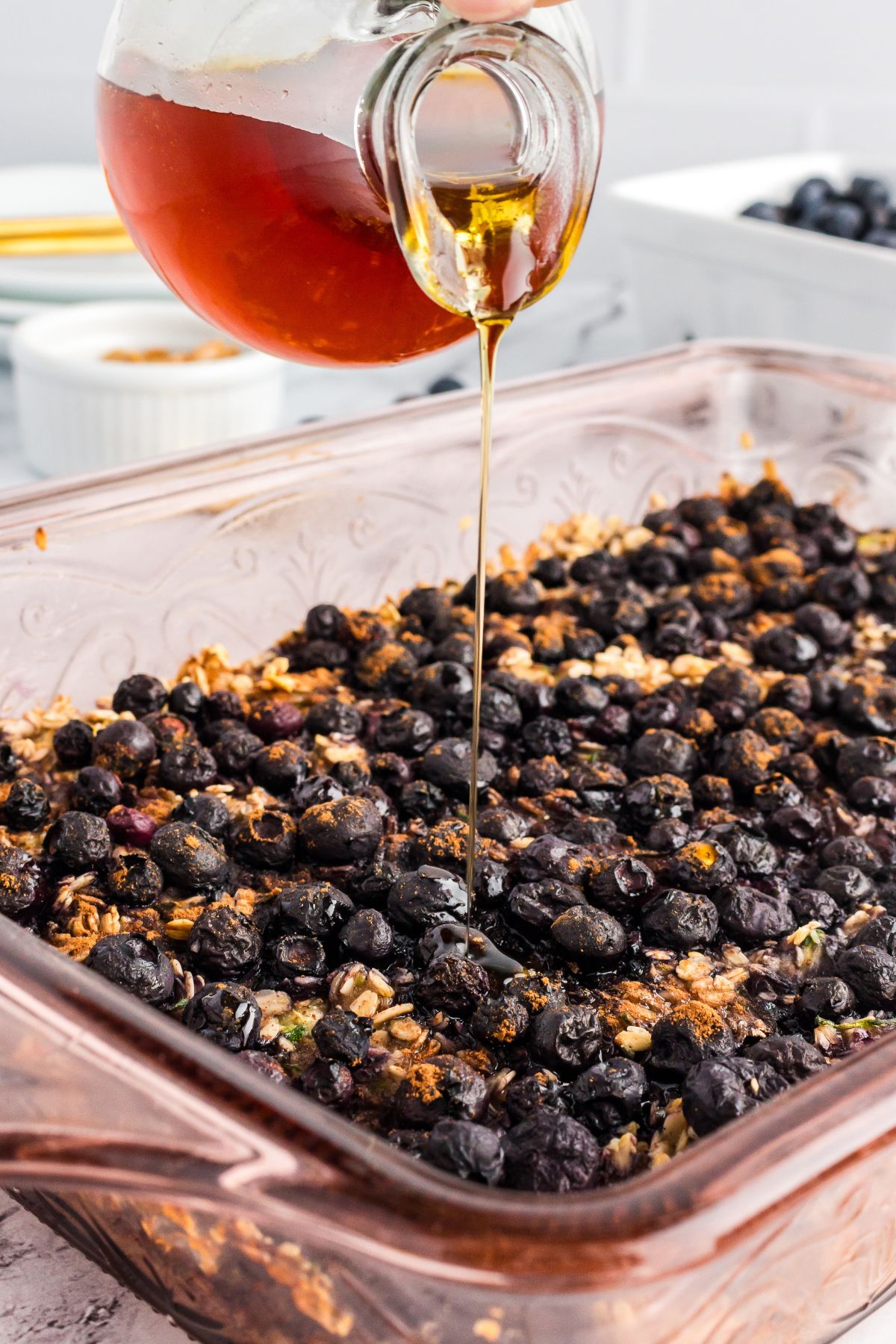 A glass jug pouring maple syrup over a freshly baked dish of zucchini blueberry oatmeal.