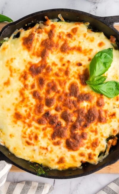 A cast iron skillet filled with freshly baked zucchini lasagna topped with browned cheese and garnished with basil, surrounded by plates, cutlery, and Parmesan.