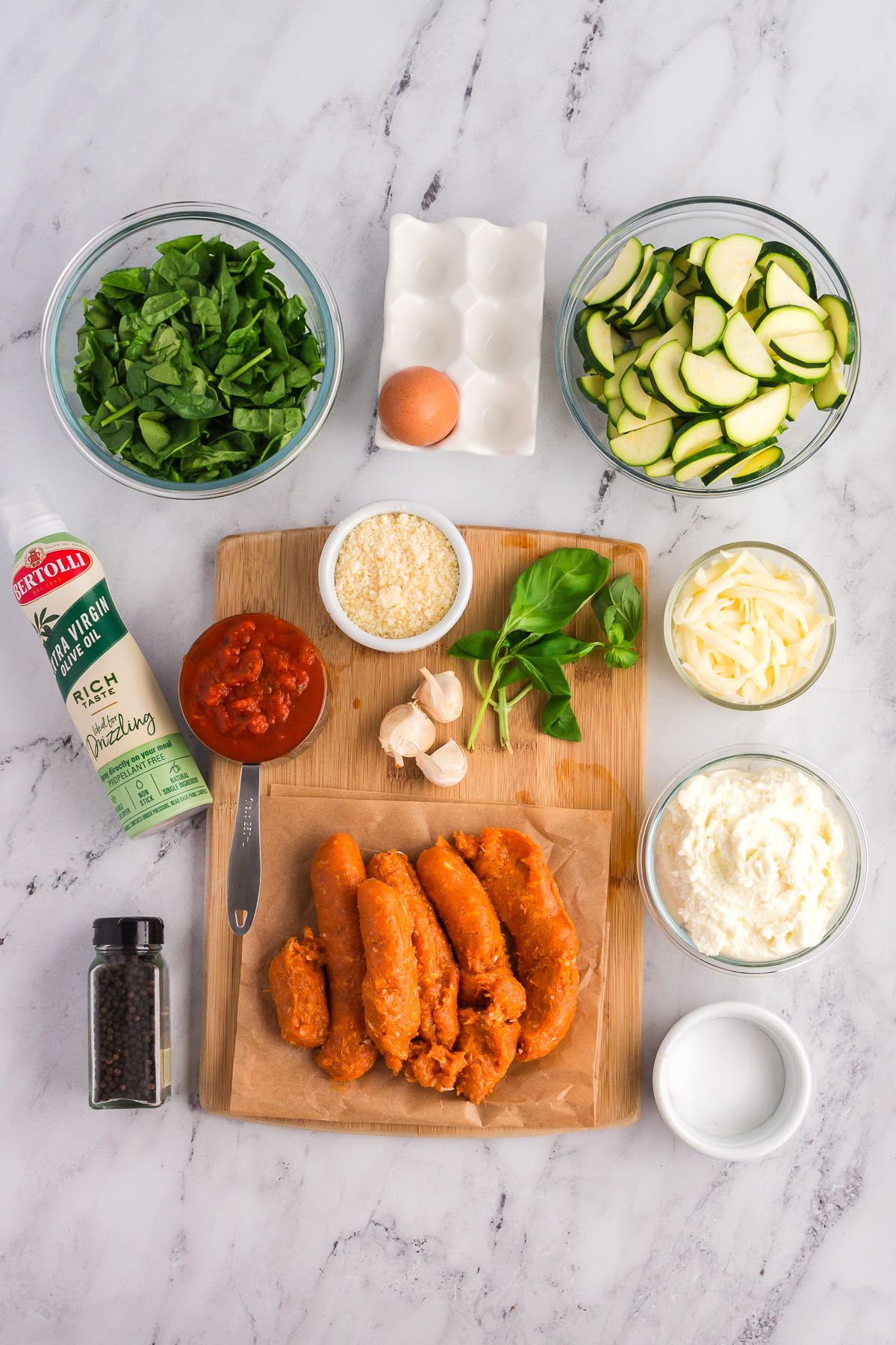 Ingredients for zucchini lasagna, including spinach, zucchini, egg, ricotta, mozzarella, tomato sauce, Parmesan, garlic, sausage, basil, olive oil spray, and black pepper, arranged on a marble countertop.