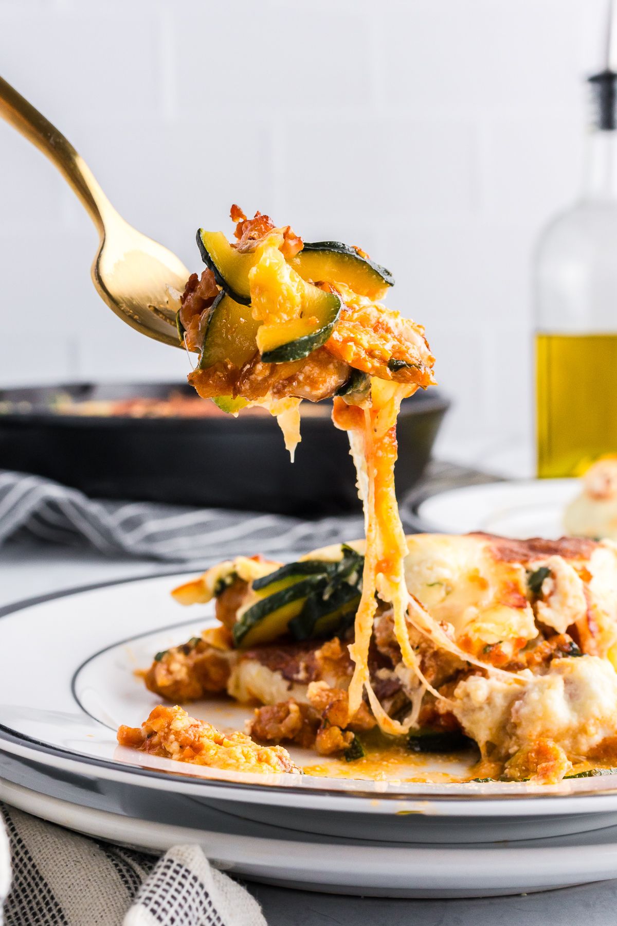 A close-up of a forkful of zucchini lasagna with layers of zucchini, melted cheese, sausage, and sauce, with a pan of the dish in the background.