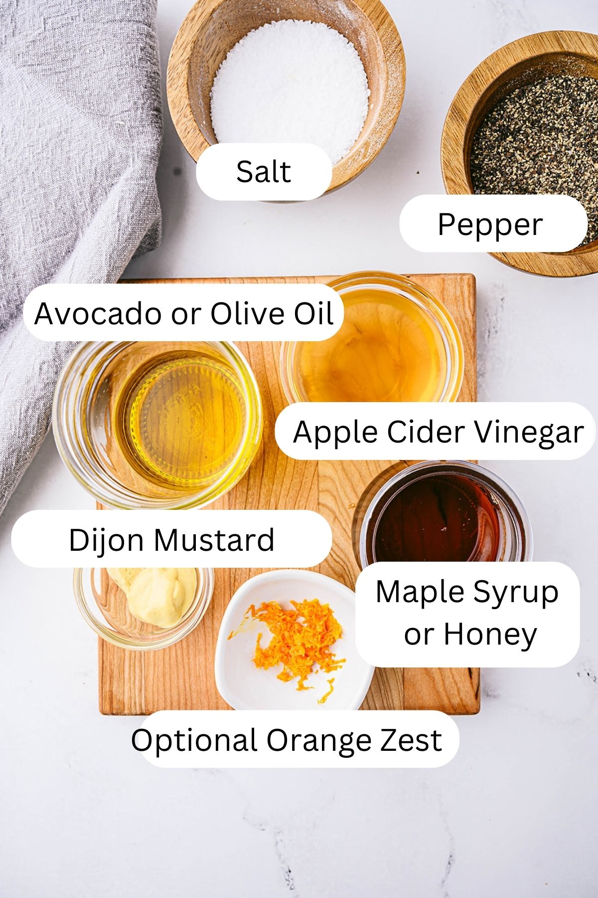 Ingredients for apple cider vinaigrette on a wooden board, including bowls of salt, pepper, avocado or olive oil, apple cider vinegar, Dijon mustard, maple syrup or honey, and optional orange zest.