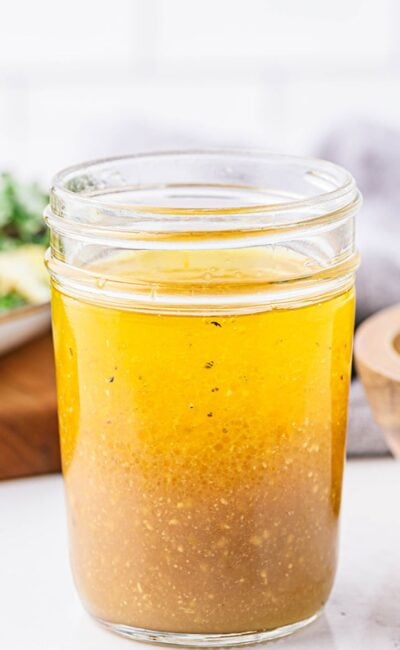 A jar of freshly mixed apple cider vinaigrette, with the layers of oil and vinegar starting to combine. A salad and wooden bowl with pepper are visible in the background.