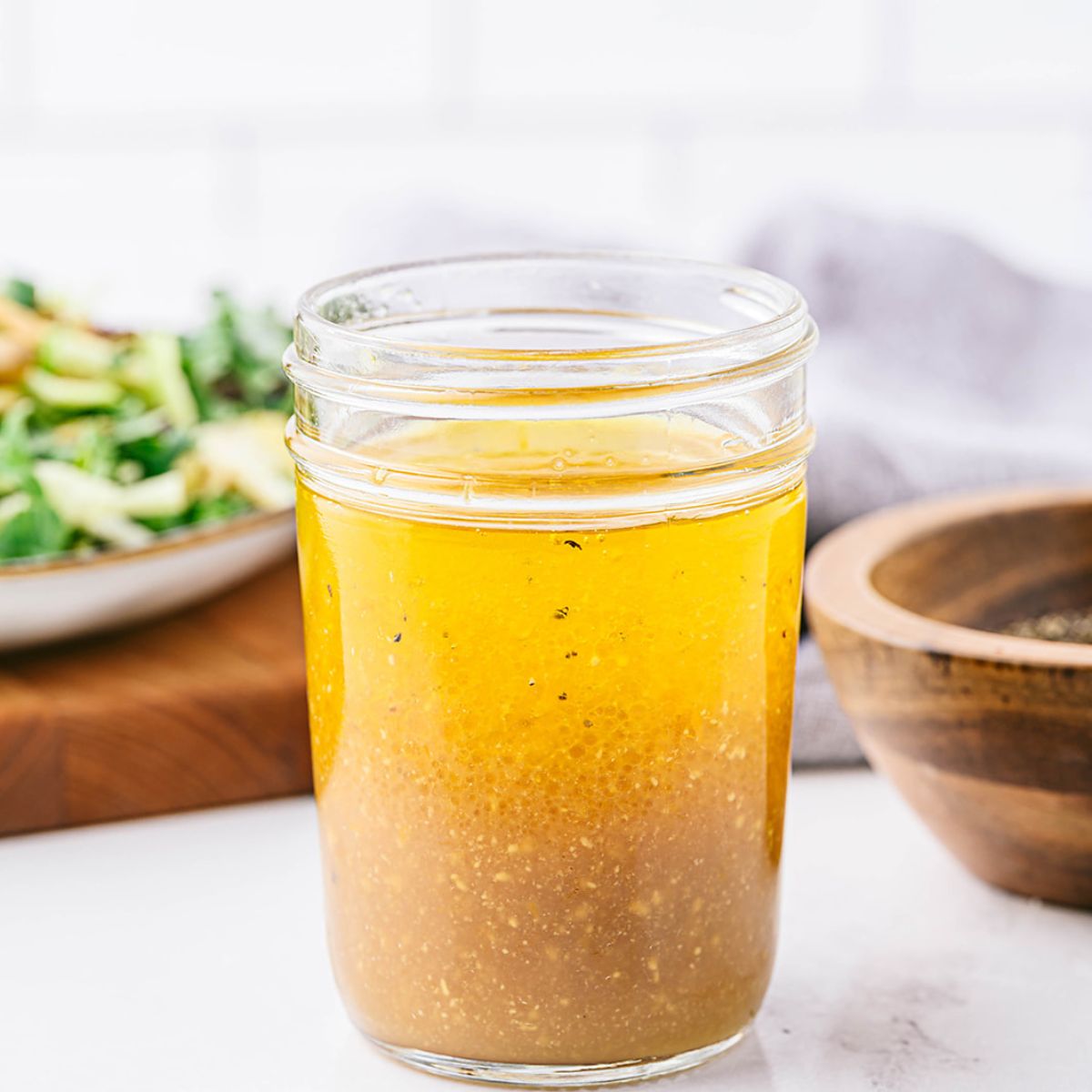 A jar of freshly mixed apple cider vinaigrette, with the layers of oil and vinegar starting to combine. A salad and wooden bowl with pepper are visible in the background.