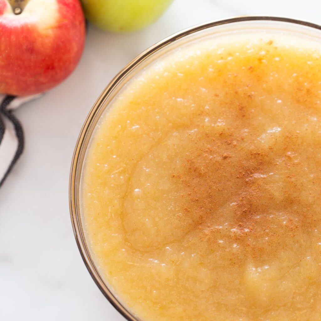 A clear bowl of applesauce with apples in the upper left hand corner 