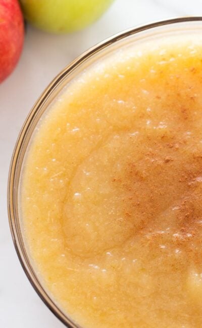 A clear bowl of applesauce with apples in the upper left hand corner