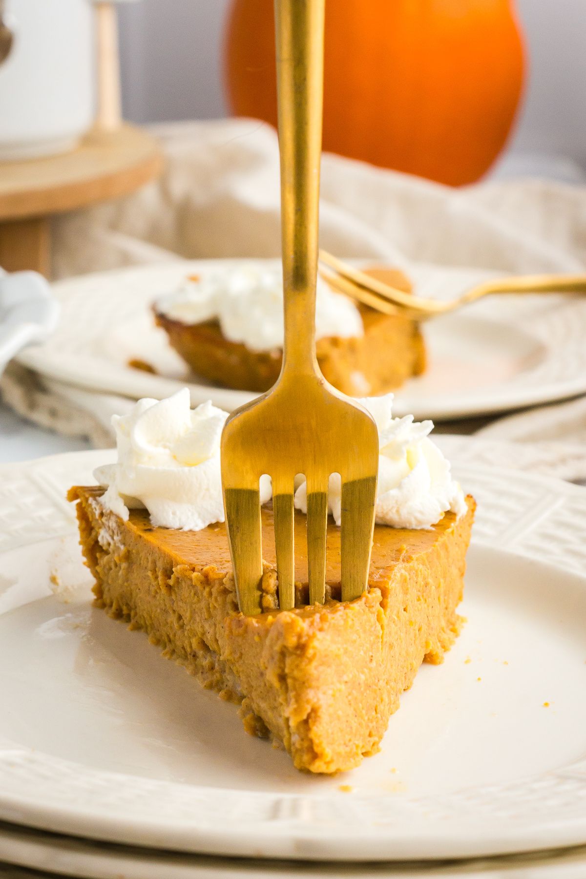 A close-up of a slice of crustless pumpkin pie on a white plate, topped with whipped cream, with a gold fork standing upright in the center.