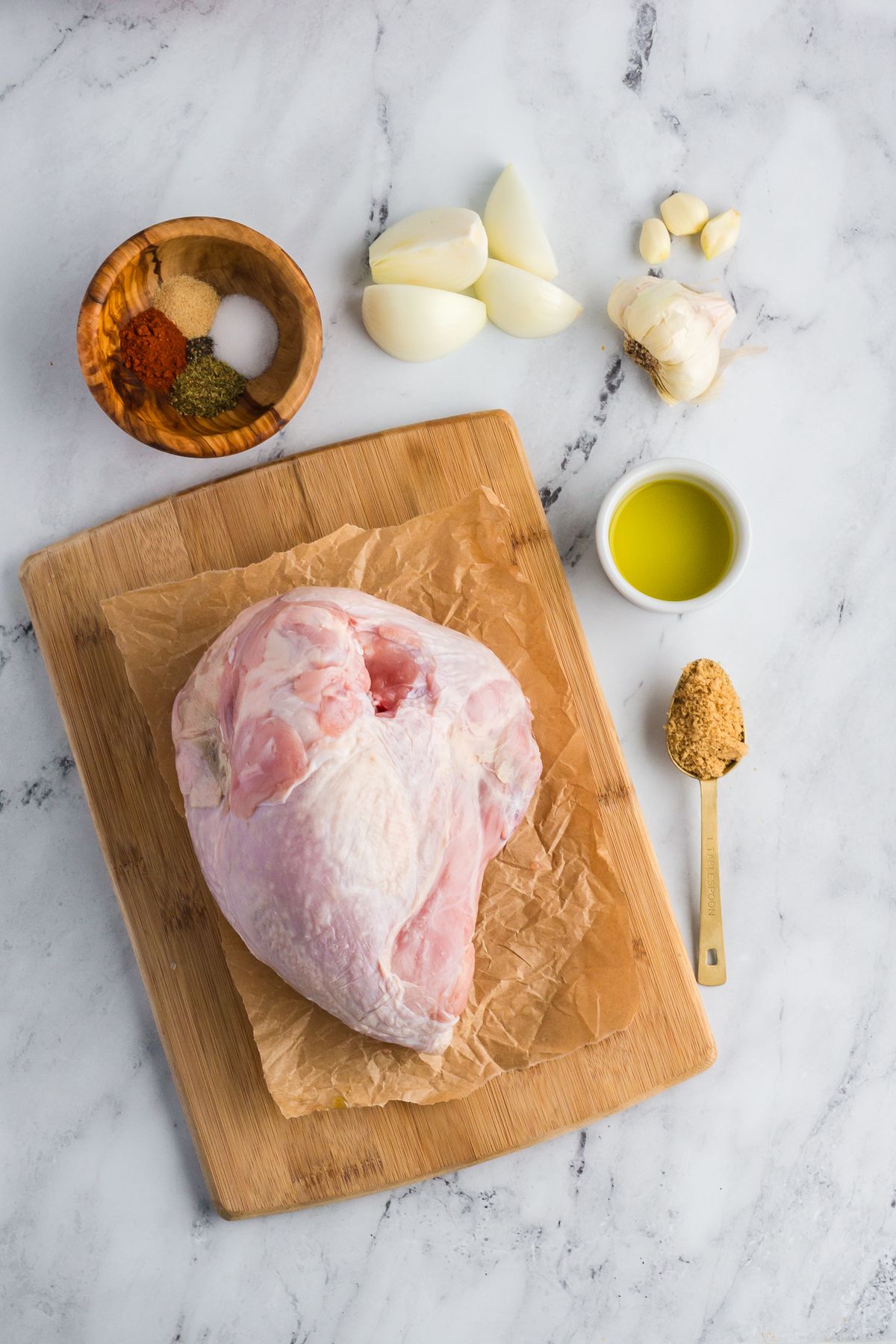 Overhead shot of ingredients for slow cooker turkey breast arranged neatly on a countertop and cutting board, including olive oil, brown sugar, spices, salt, pepper, onion, and garlic.