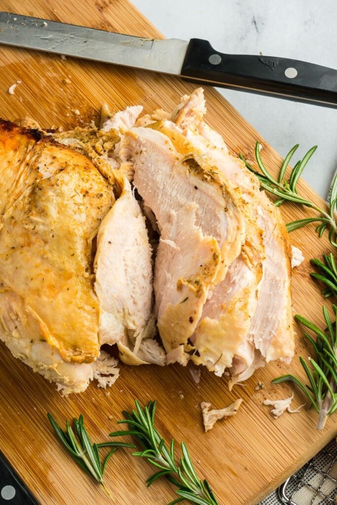 Sliced turkey breast on a wooden cutting board, showing tender, juicy meat with golden-brown crispy skin and rosemary garnish.