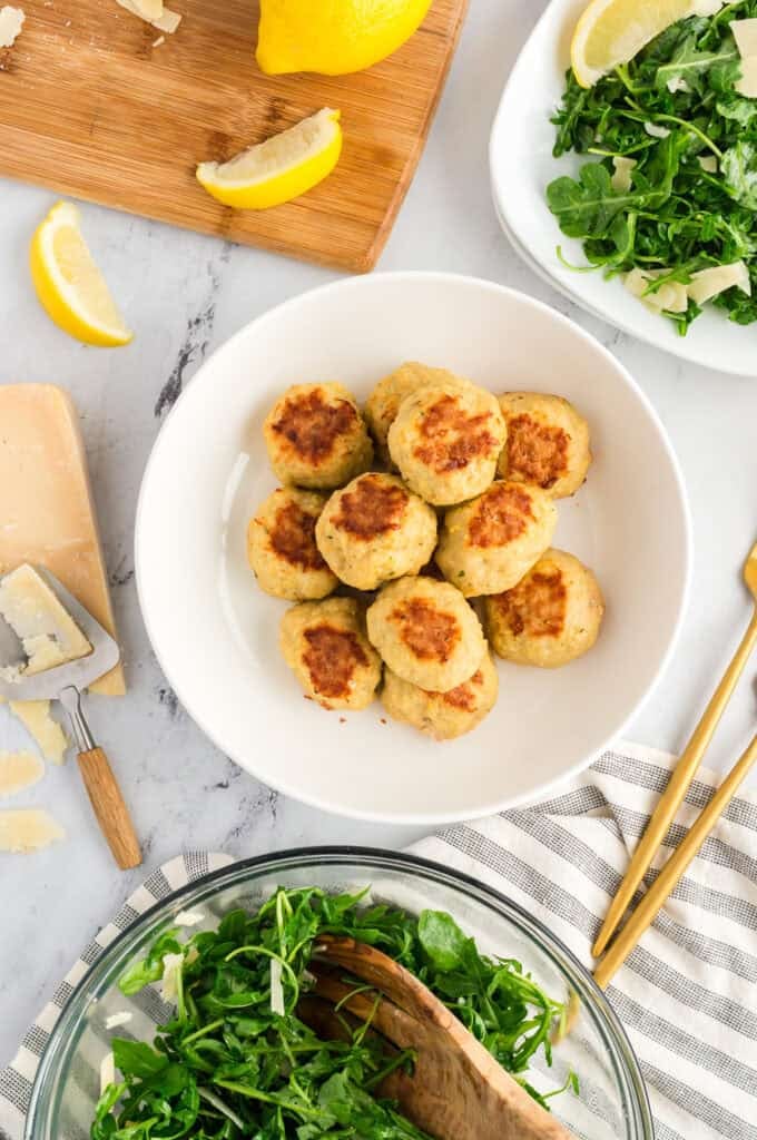 Overhead shot of chicken meatballs on a white table with a serving bowl and plate of arugula salad.