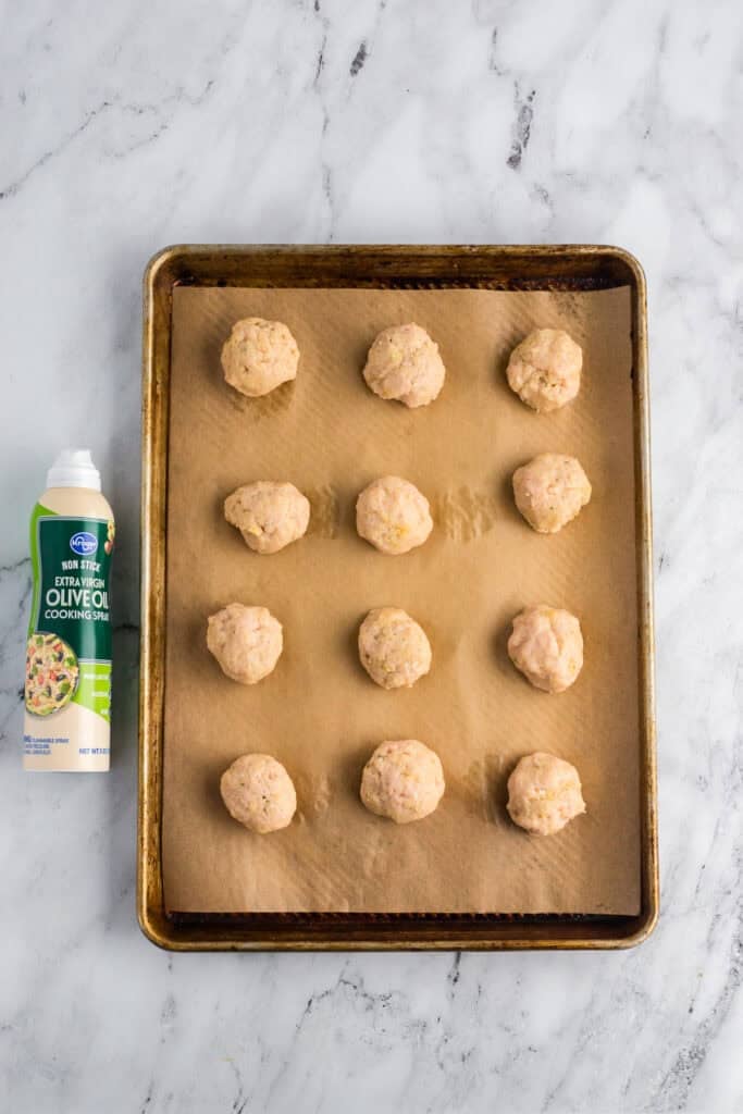 Parchment lined sheet pan with raw chicken meatballs sitting next to olive oil cooking spray.