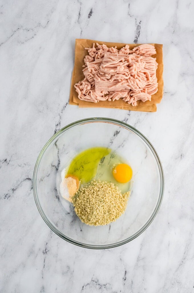 Glass bowl with eggs and breadcrumb sitting next to lean ground chicken on parchment paper.