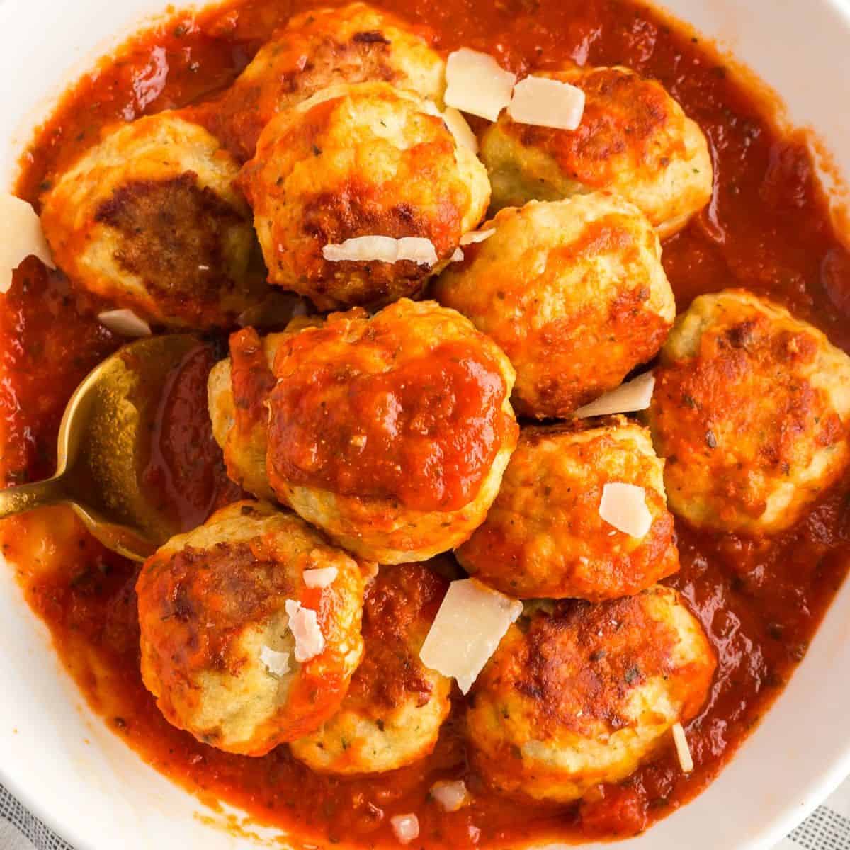 Baked chicken meatballs in a white bowl of sauce topped with shaved parmesan cheese.