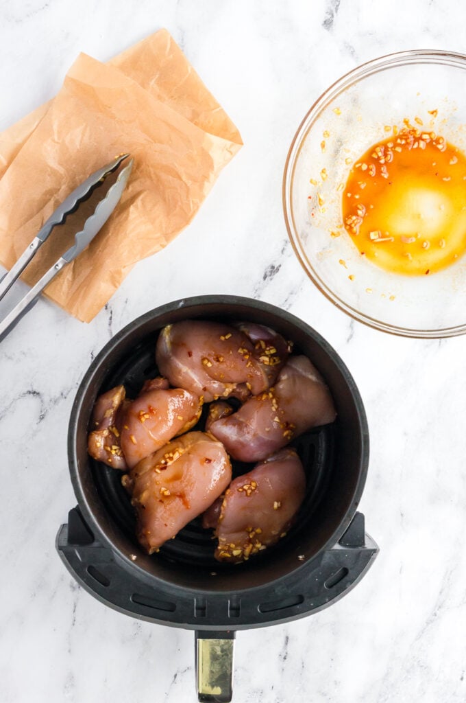 Marinated chicken thighs placed in a single layer in an air fryer basket on a white marble background.