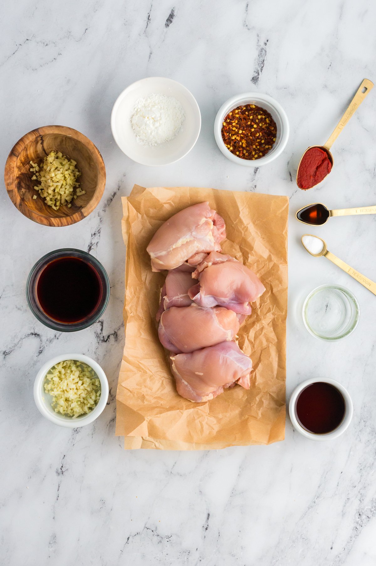 raw boneless skinless chicken thighs on parchment paper all on a marble background with all the ingredients for the marinade and sauce in separate containers around them.