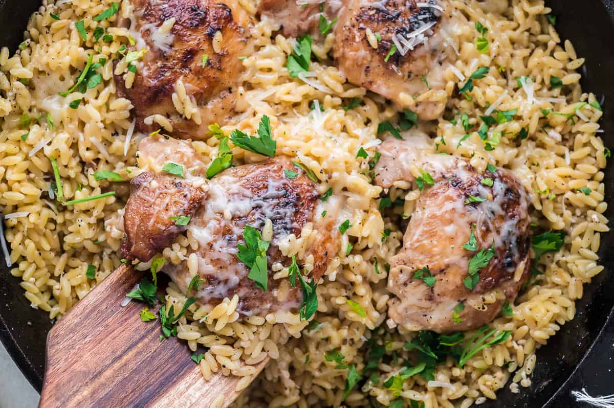 A piece of chicken thigh on a wooden spatula held over the pan of chicken and orzo.