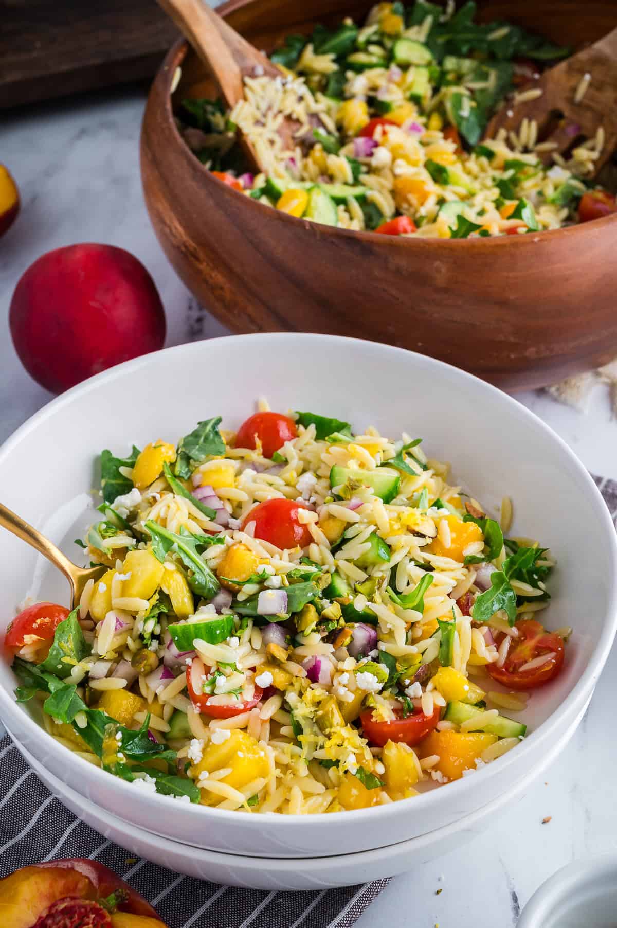 Individual serving of summer orzo salad in a white bowl beside a large wooden bowl filled with the remaining salad.